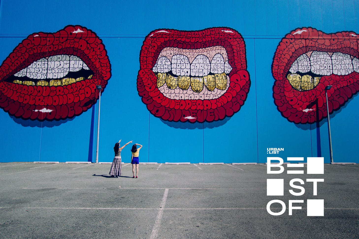 Two people gaze up at a huge street artwork showing three red mouths on a blue background.